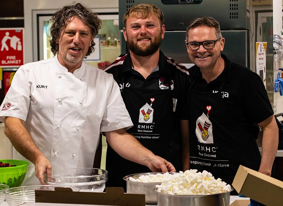 Three men smiling in the Auckland RMHC kitchen, one in chef whites, standing in front of bowls of marshmellows.