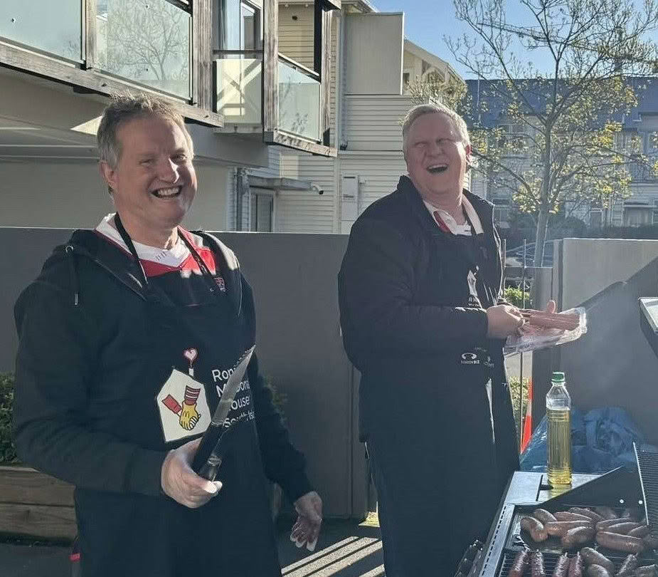 Two men outside laughing cooking sausages on the BBQ.