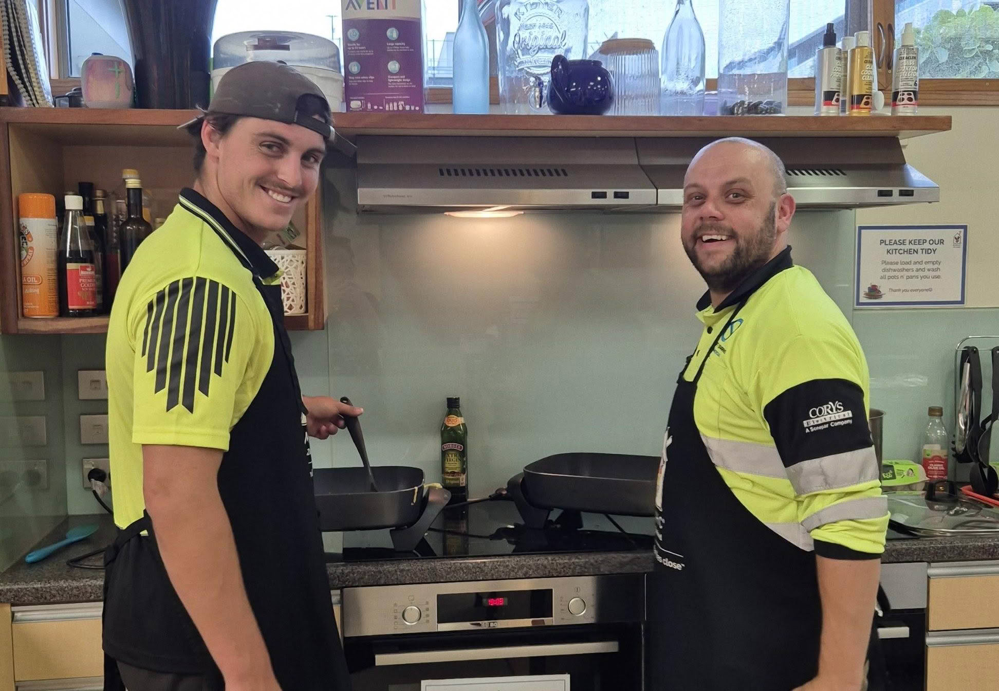 Two men turning to smile at the camera cooking dishes on the stove.