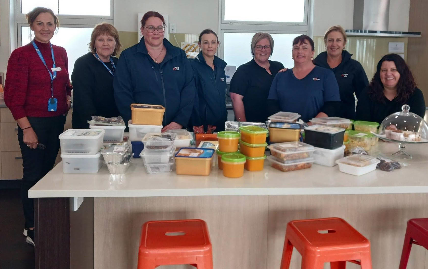 Eight women smiling at the camera standing in front of a bench filled with baking and meals in containers for families.