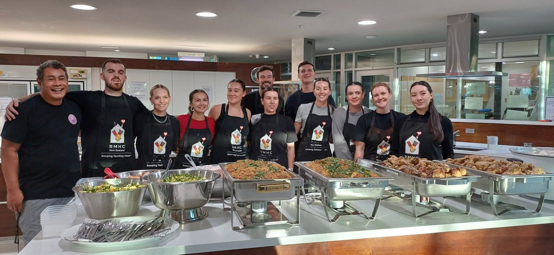 Deadly Ponies employees standing in a line and smiling in front of the meal they have cooked at RMHC's family dinner night.